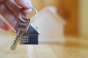 anahtar-teslim Hand holding giving key chain with house shaped pendant on blurred wooden table and home model background with copy space. Real estate, buying moving new home, renting property or apartments concept.